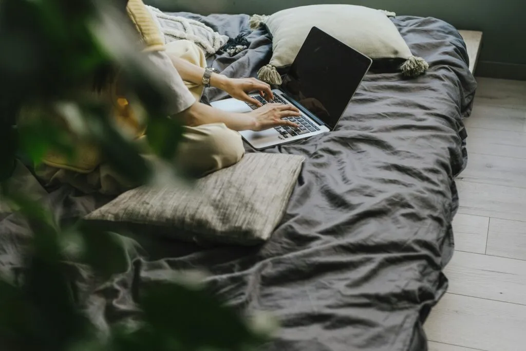 Someone working on a laptop at home, sitting on a bed with cushions