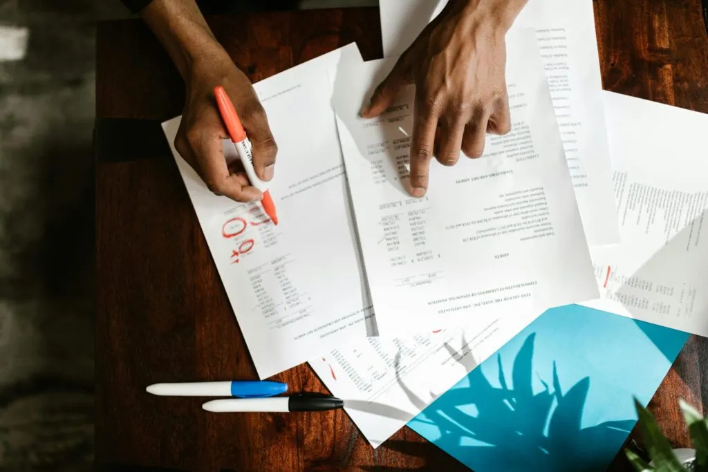 Printed financial schedules being checked and annotated with a red pen on a desk