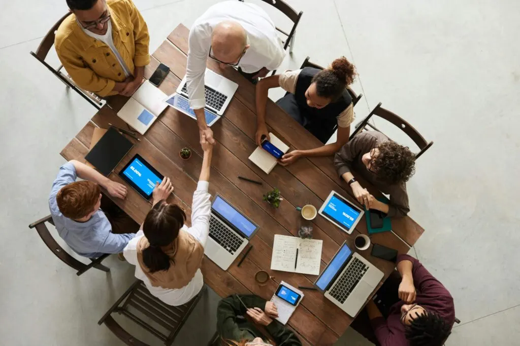 Overhead view of a business team around a wooden table with laptops and tablets, two of them shaking hands